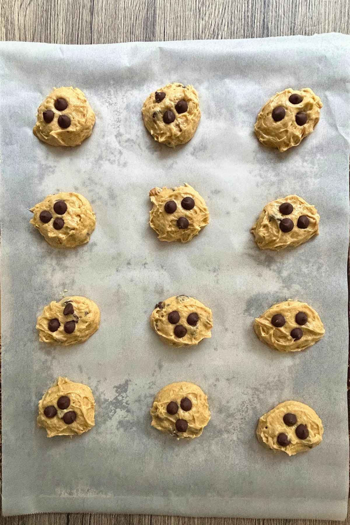 Raw scoops of cookie dough studded with chocolate chips arranged on a parchment-lined baking sheet.