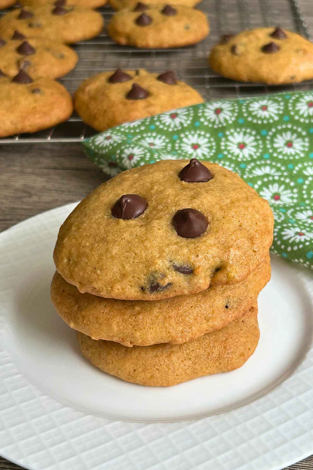 Stack of three golden-brown banana chocolate chip cookies on a white textured plate next to a green patterned napkin.