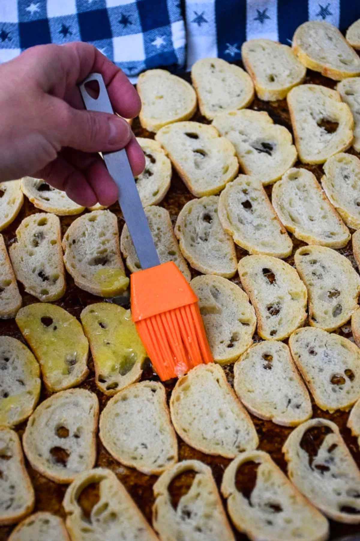 A hand holding an orange silicone brush spreading garlic butter overtop of baguette slices on a baking sheet.