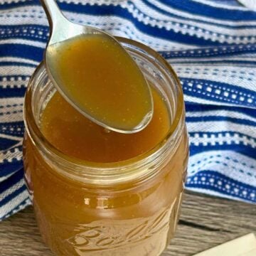 A silver spoon holding rich, golden butterscotch sauce over an open Ball Mason jar with a vintage handwritten recipe card reading "Butterscotch Sauce."