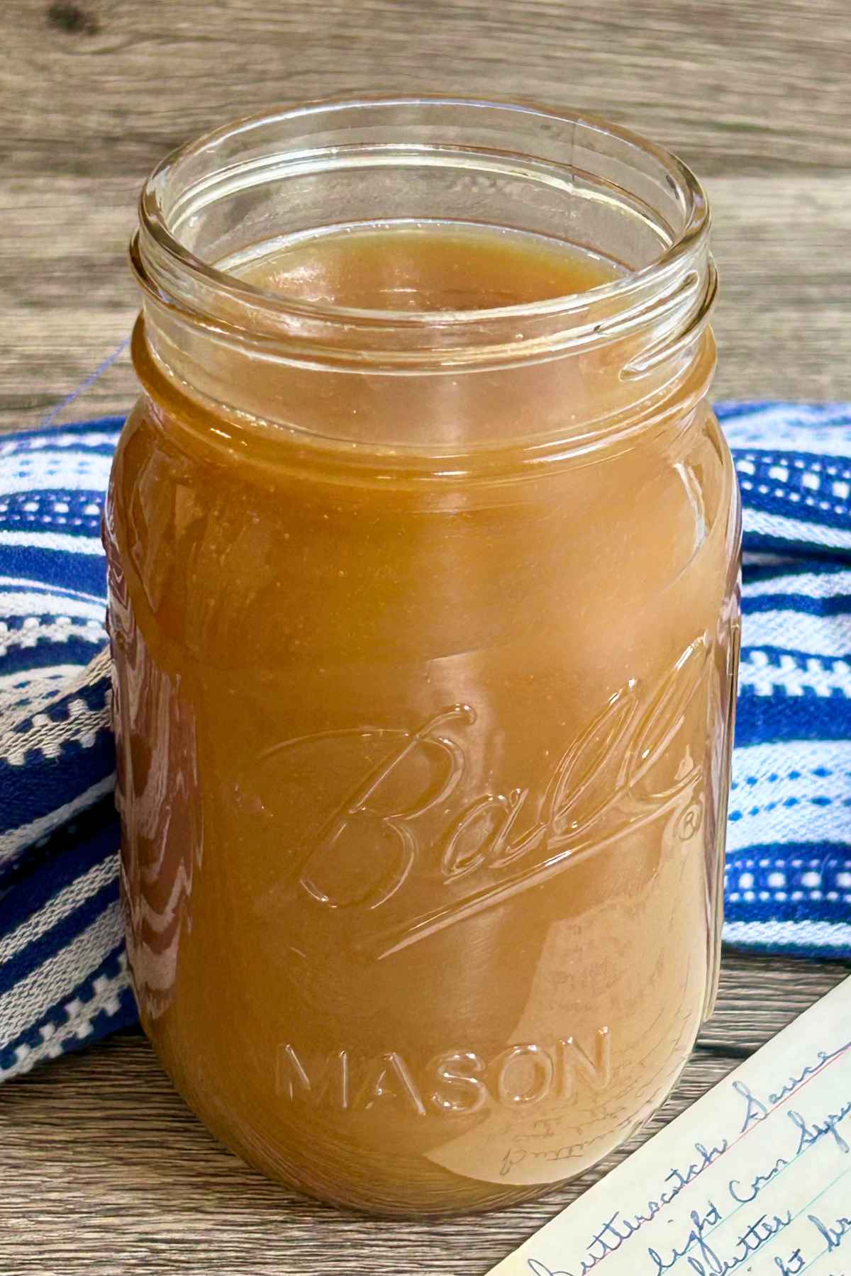A Ball mason jar filled with warm butterscotch sauce sitting on a blue and white plaid towel next to a vintage handwritten recipe card.