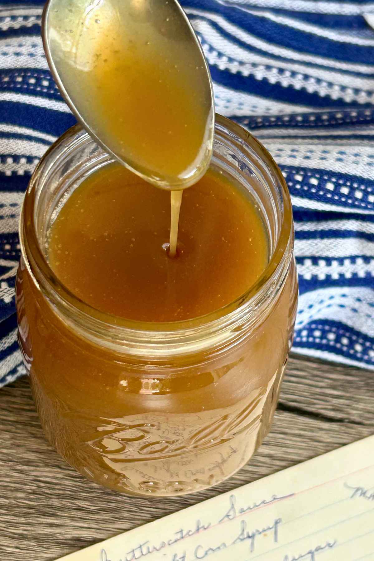 A photograph showing a close-up of a glass jar with golden-brown butterscotch sauce and a silver spoon, with a hand-written recipe card beneath the jar and a blue and white towel to the side.