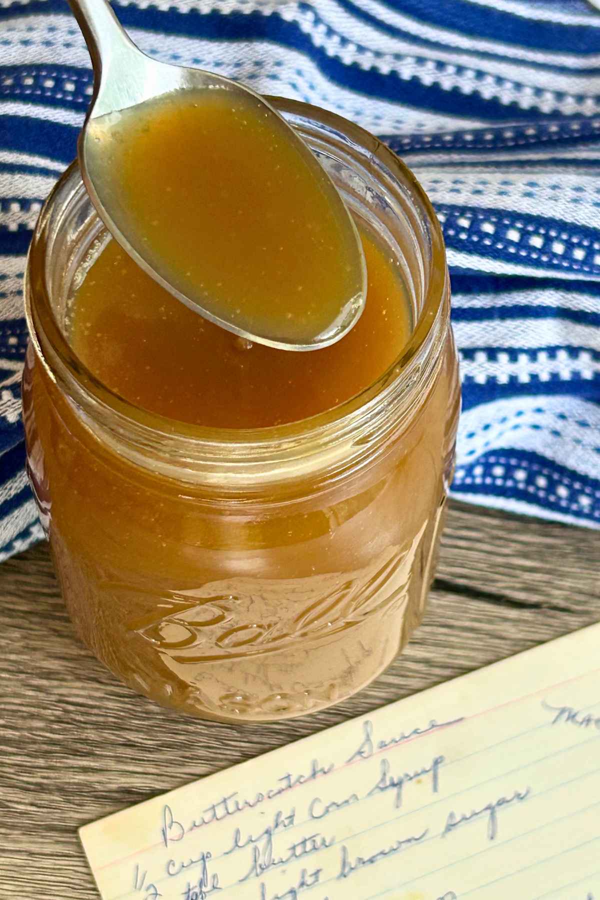 A silver spoon holding rich, golden butterscotch sauce over an open Ball Mason jar with a vintage handwritten recipe card reading "Butterscotch Sauce."