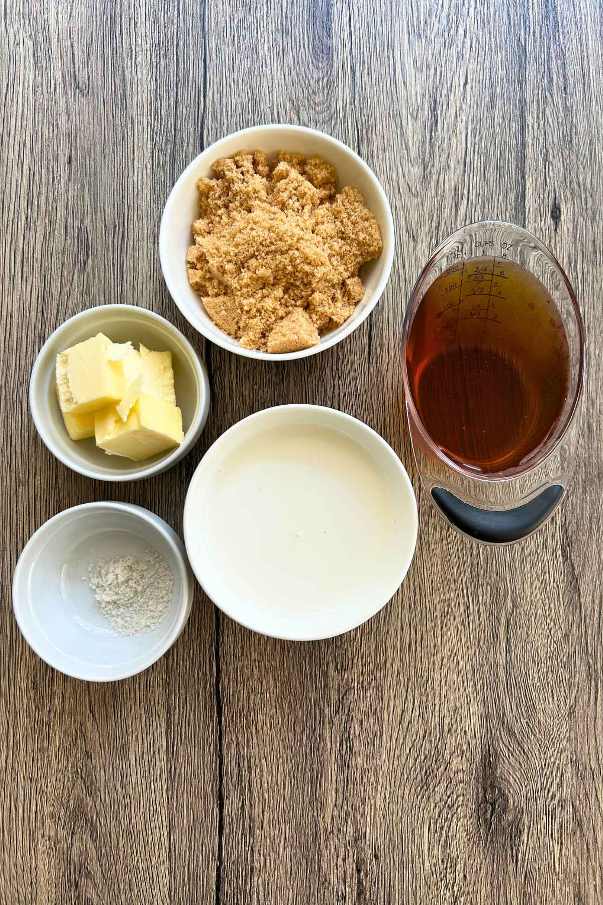 Ingredients to make homemade sauce, including brown sugar, butter, heavy cream, salt, and maple syrup arranged in small bowls on a wooden surface.