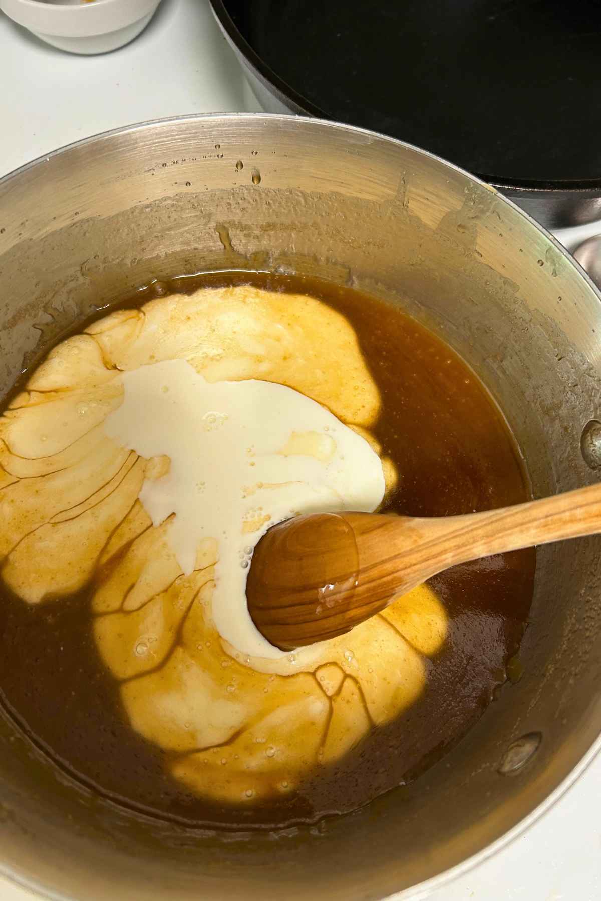 A saucepan filled with melted brown sugar and maple syrup mixture topped with cream, ready to stir with a wooden spoon.