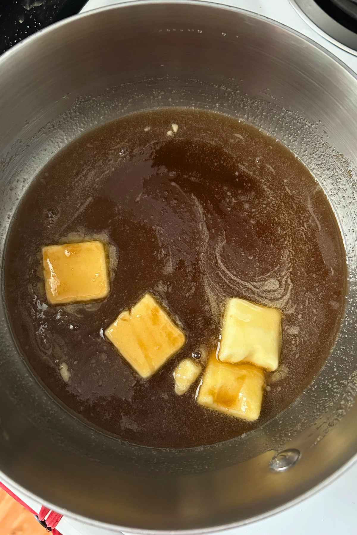 Pats of butter melting into brown sugar and maple syrup inside a stainless steel saucepan.