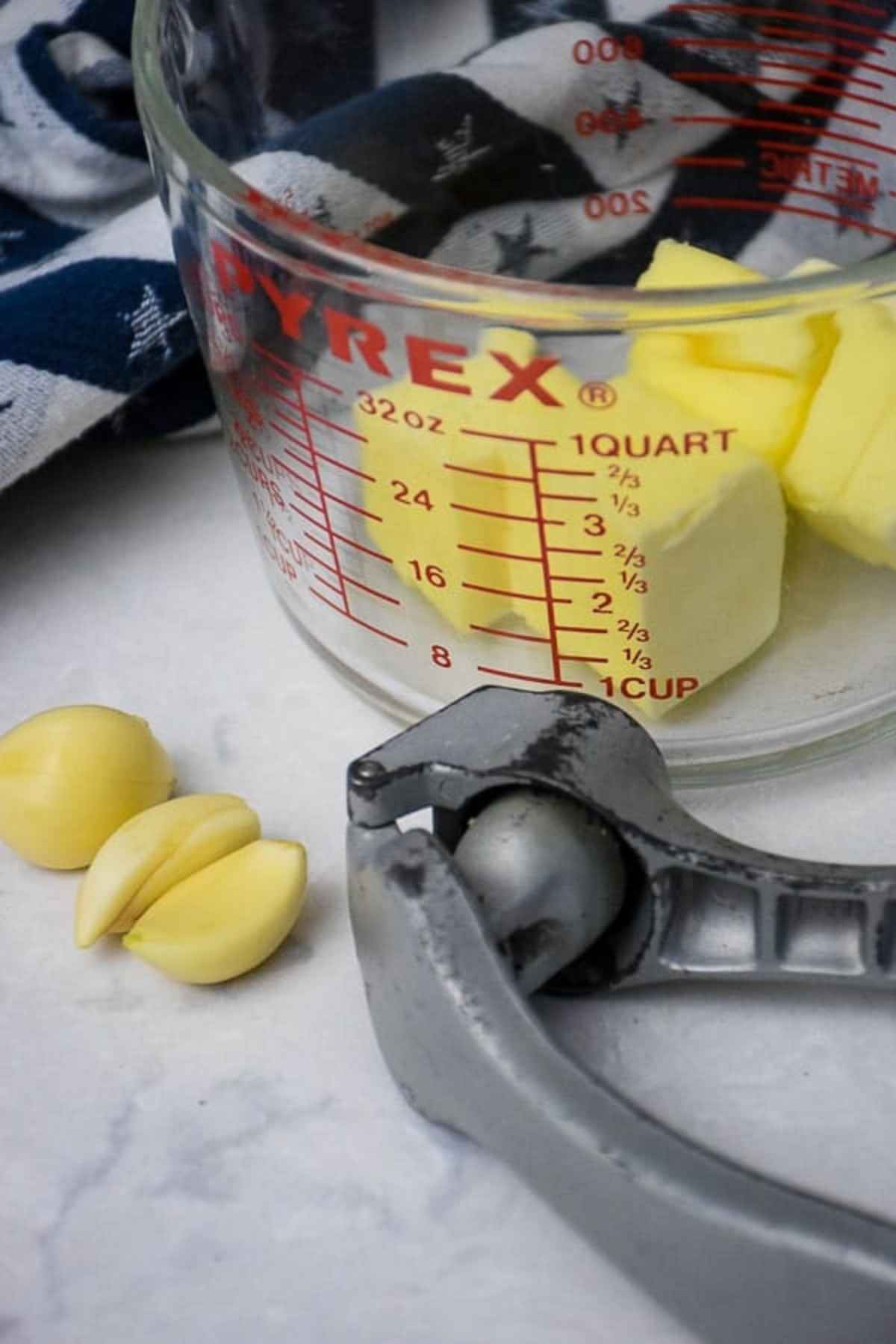 A glass measuring cup with cubed butter inside with a silver garlic press and garlic cloves in front of the measuring cup.