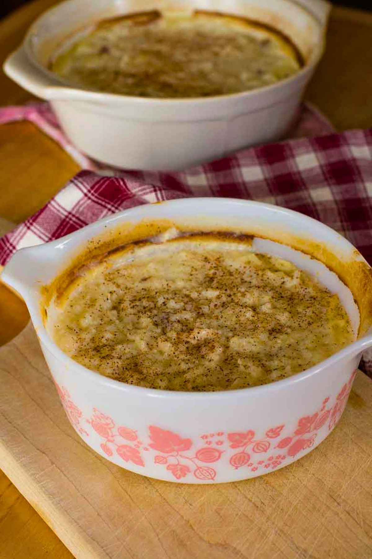 Two dishes of baked rice pudding with a golden custard top, one in a vintage white and pink Pyrex bowl, set on a wooden cutting board with a red plaid kitchen towel.