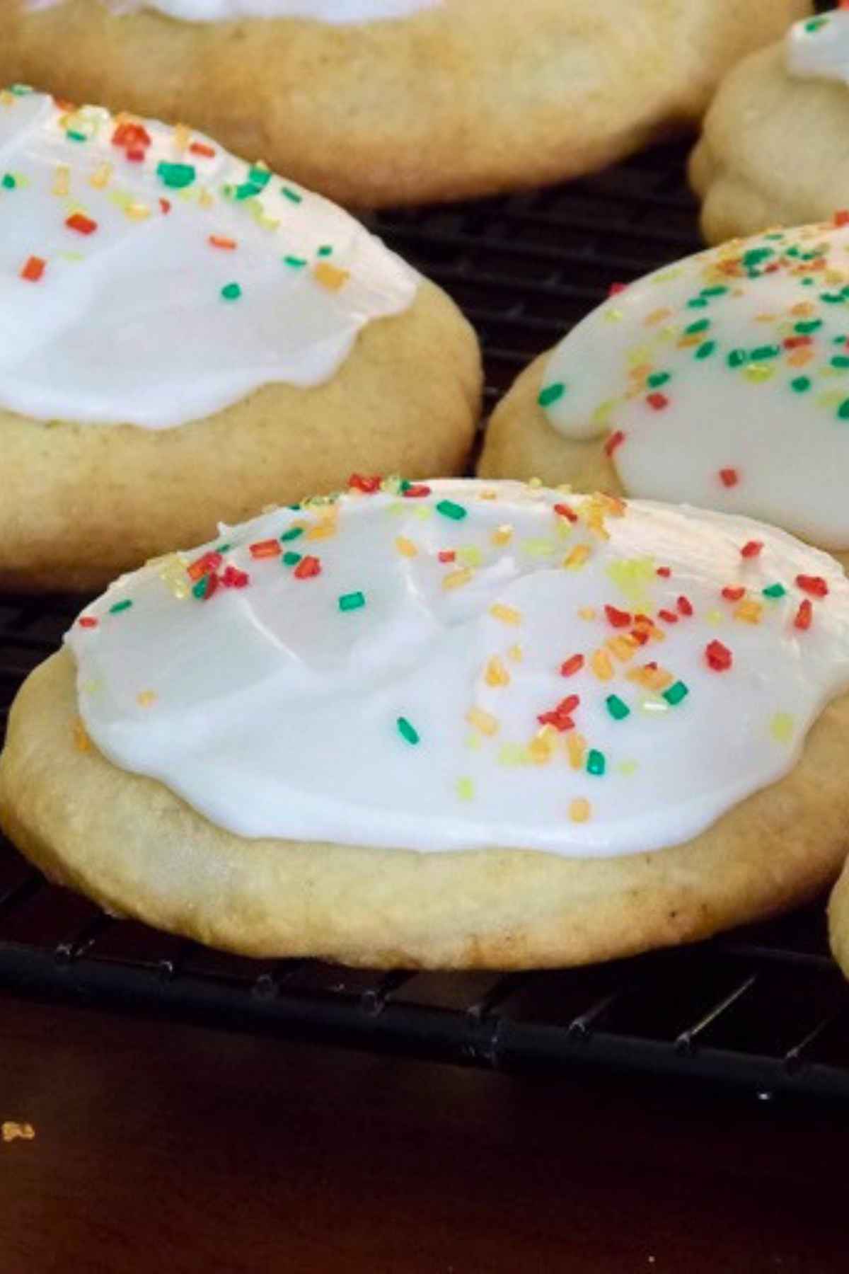 A close-up of soft frosted cookies topped with colorful sprinkles cooling on a black wire rack.