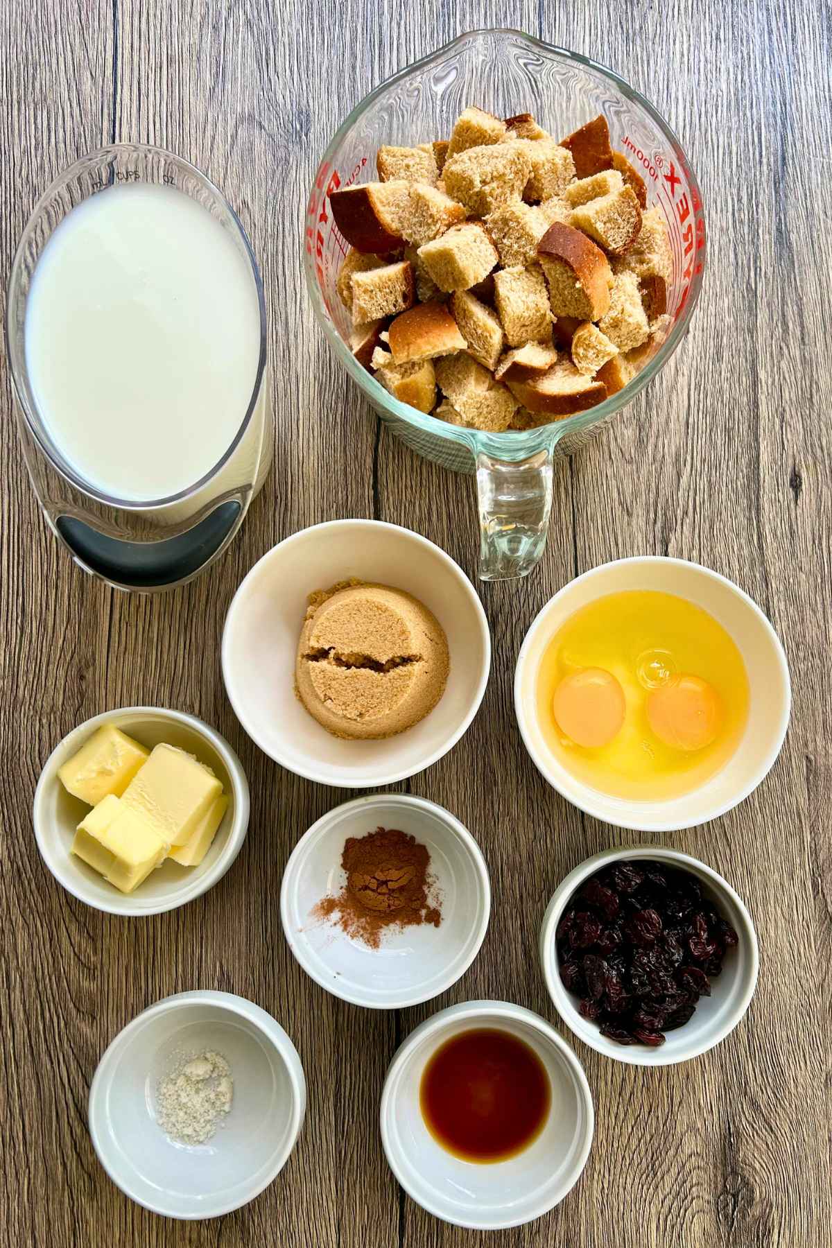 An overhead view of ingredients arranged in small white bowls and glass measuring cups on a wooden surface, including milk, cubed bread, brown sugar, eggs, butter, raisins, spices, flour, and vanilla extract.