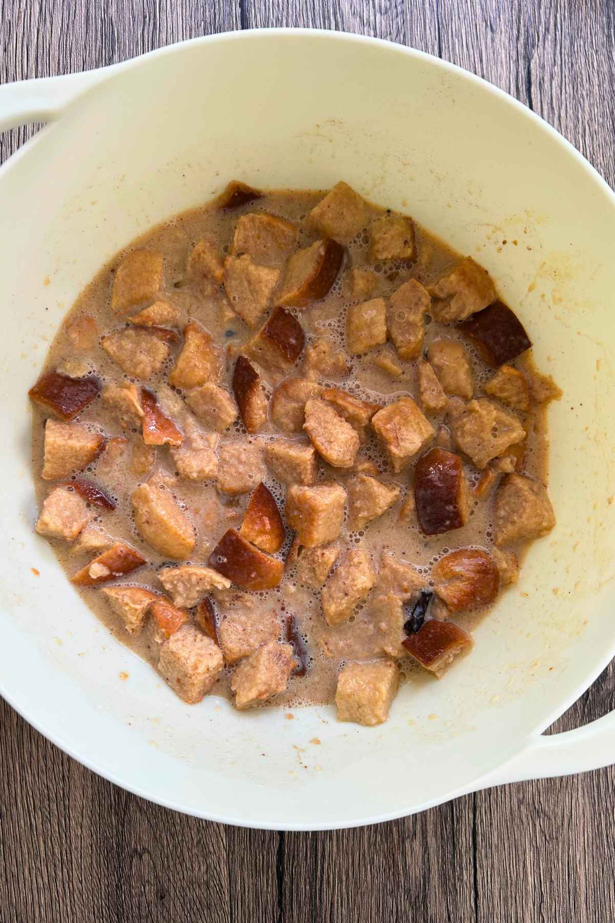 An overhead view of cubed bread soaking in a creamy brown liquid mixture inside a large white bowl on a wooden surface.
