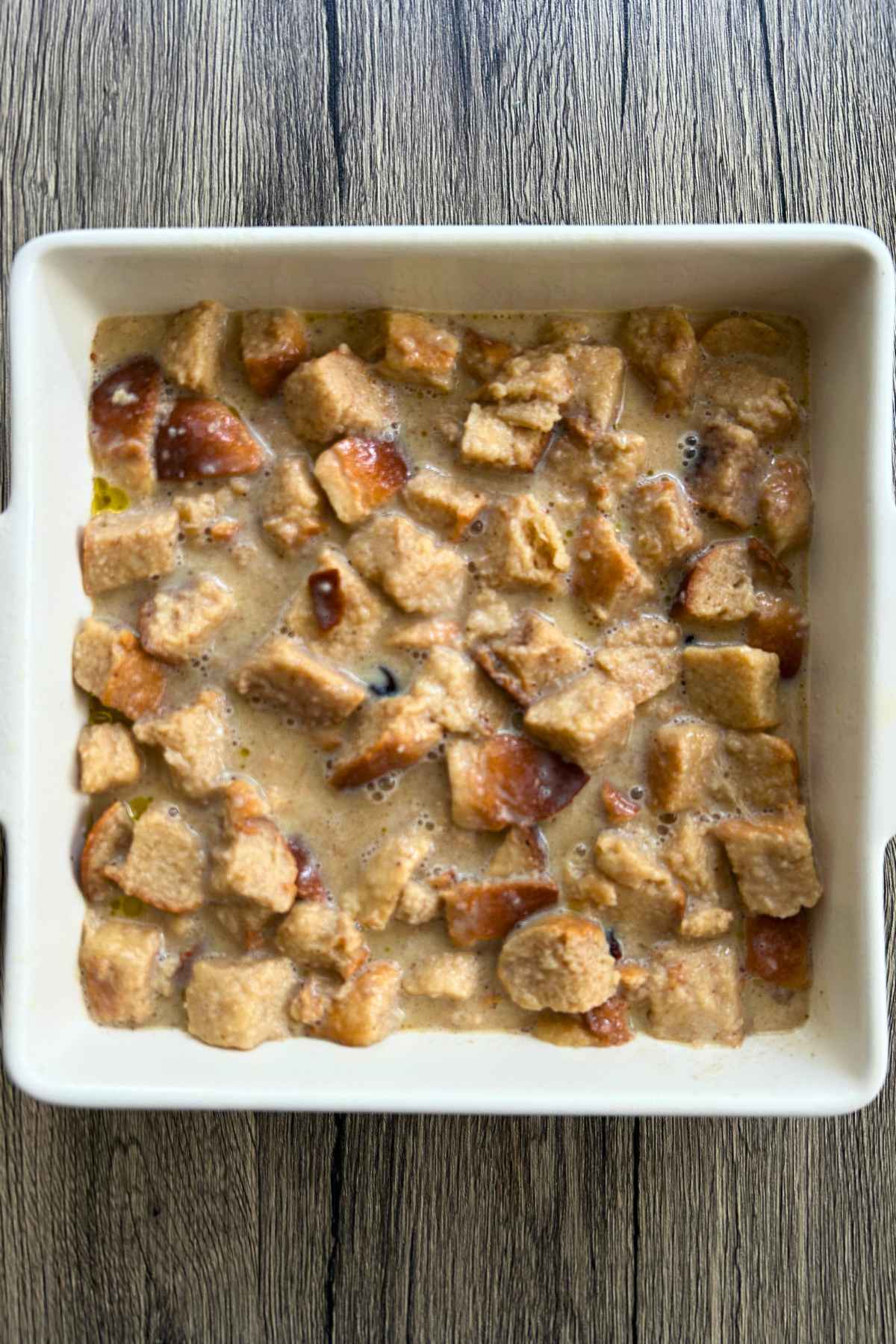 An overhead view of cubed bread soaked in a liquid mixture inside a square white ceramic baking dish before baking.