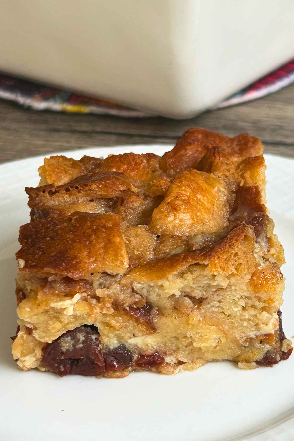 A square slice of bread pudding with a golden-brown top and visible raisins, served on a white plate with a white baking dish blurred in the background.