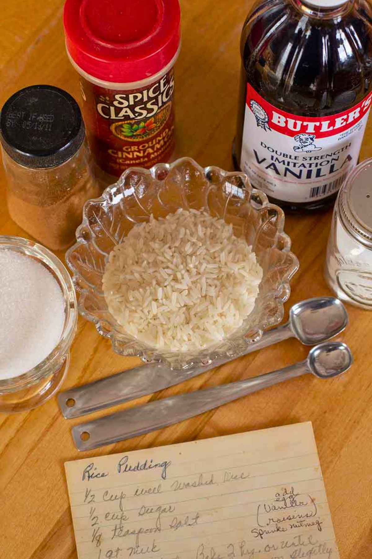 Ingredients for baked rice pudding including long grain rice, vanilla extract, cinnamon, sugar, salt, and measuring spoons arranged on a wooden surface beside a handwritten recipe card.
