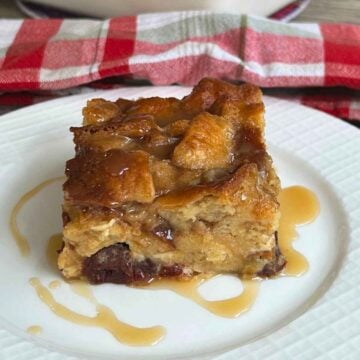 A square slice of warm old-fashioned bread pudding with raisins, drizzled with a butterscotch sauce, served on a white plate in front of a white baking dish on a red and white checkered cloth.