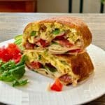 Two halves of a toasted turkey bacon breakfast burrito stacked on a white plate served with a side of fresh greens, diced tomatoes, and sliced avocado.