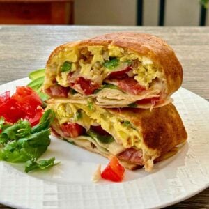 Two halves of a toasted turkey bacon breakfast burrito stacked on a white plate served with a side of fresh greens, diced tomatoes, and sliced avocado.