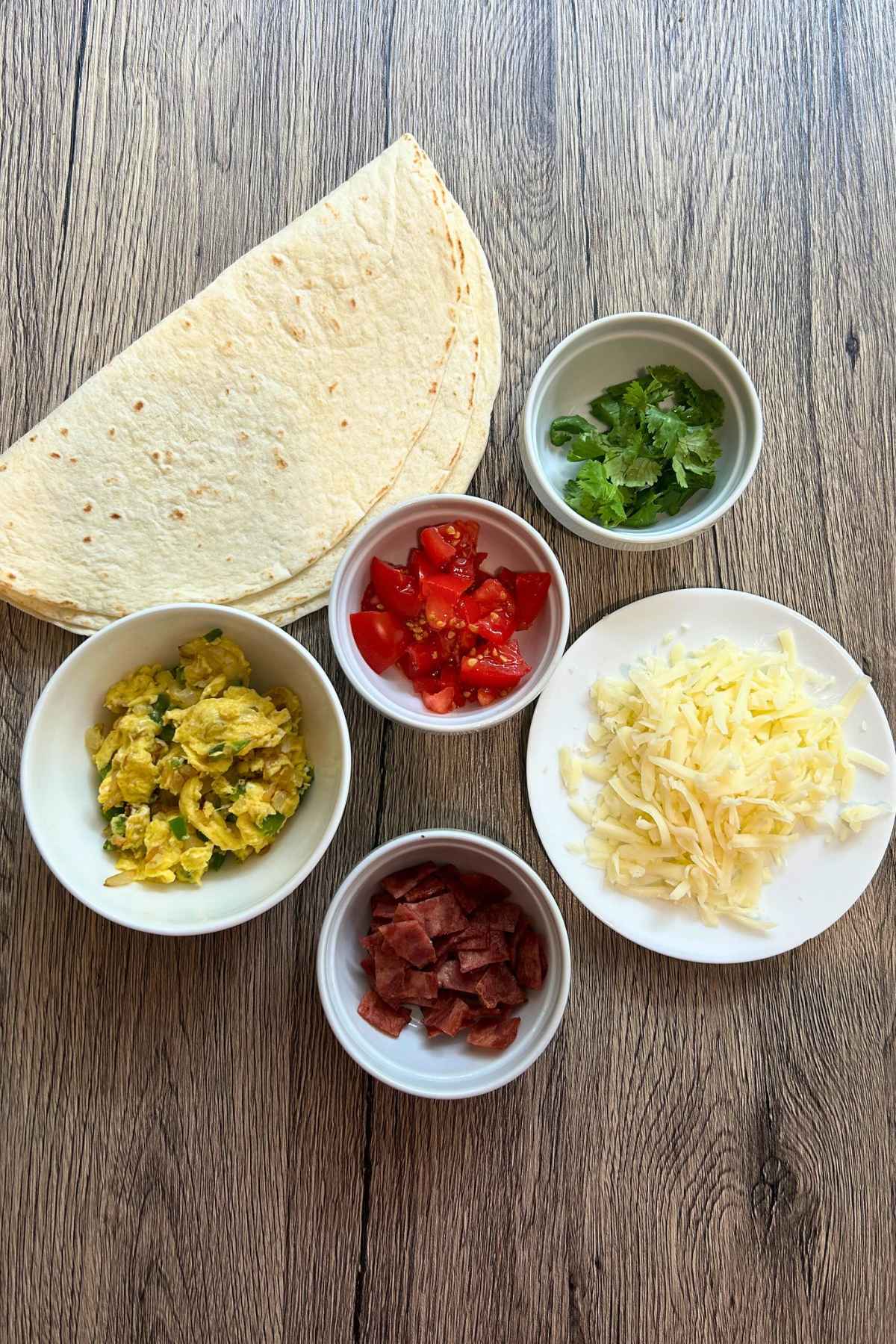 Small white bowls of scrambled eggs, diced tomatoes, chopped bacon, shredded cheese, and fresh cilantro arranged on a wooden surface next to flour tortillas.