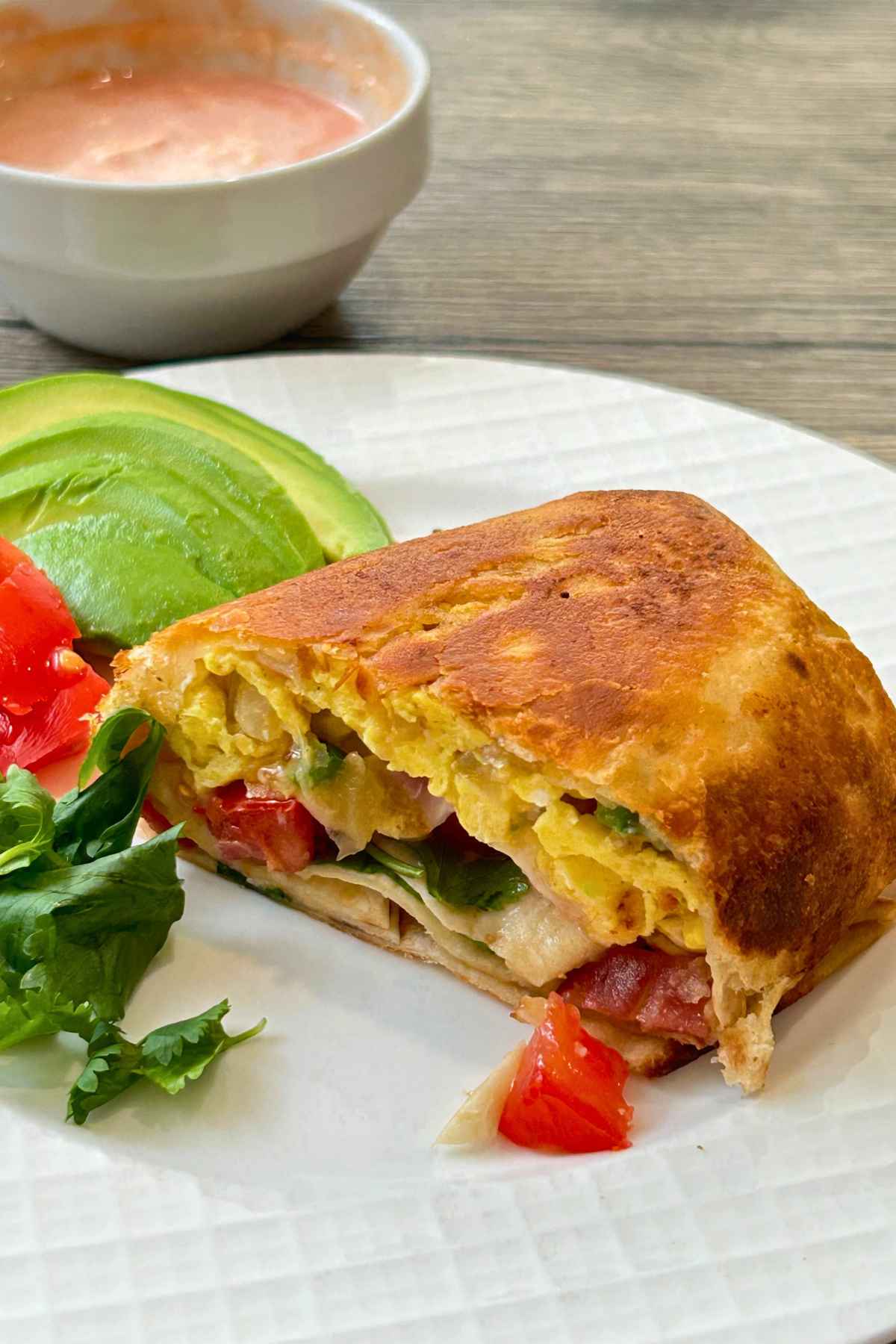 Golden-brown crispy breakfast burrito cut in half on a white plate with sliced avocado, tomatoes, cilantro, and a bowl of salsa.