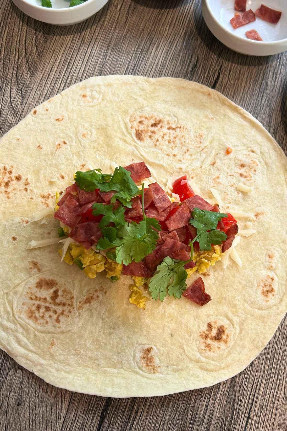 Large flour tortilla laid flat and topped with scrambled eggs, shredded cheese, topped bacon, diced tomatoes, and fresh cilantro.