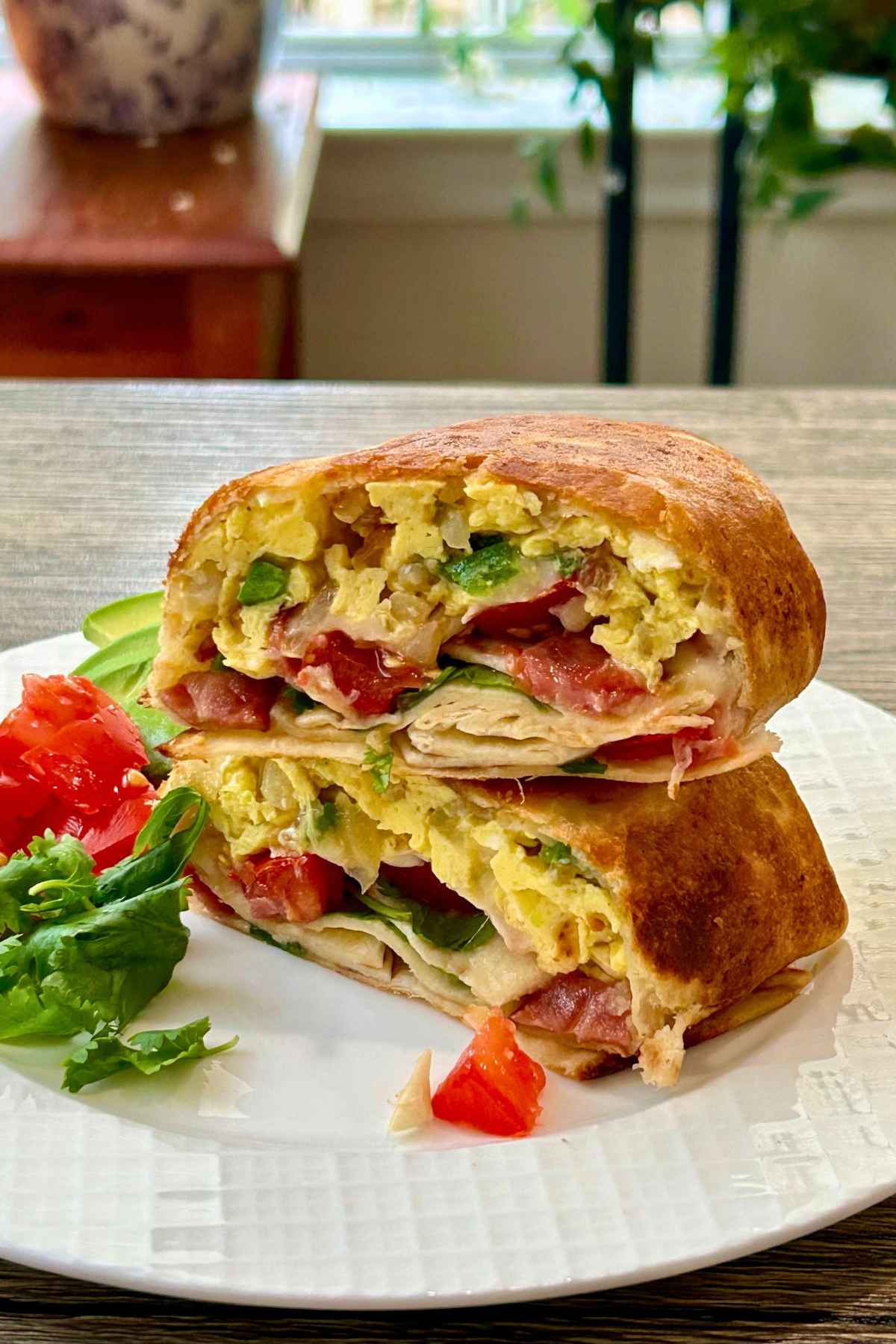 Two halves of a toasted turkey bacon breakfast burrito stacked on a white plate served with a side of fresh greens, diced tomatoes, and sliced avocado.