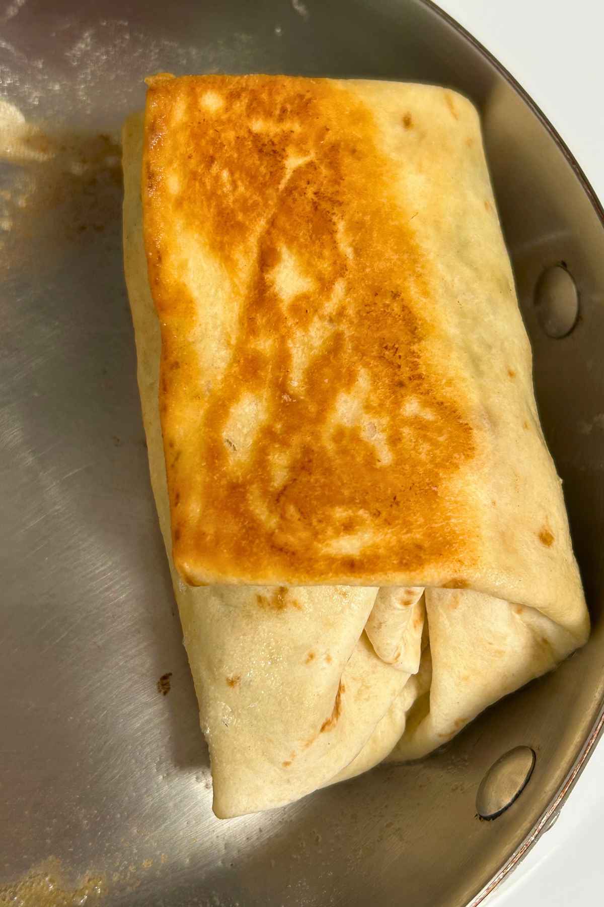 Golden-brown folded burrito searing in stainless steel skillet.