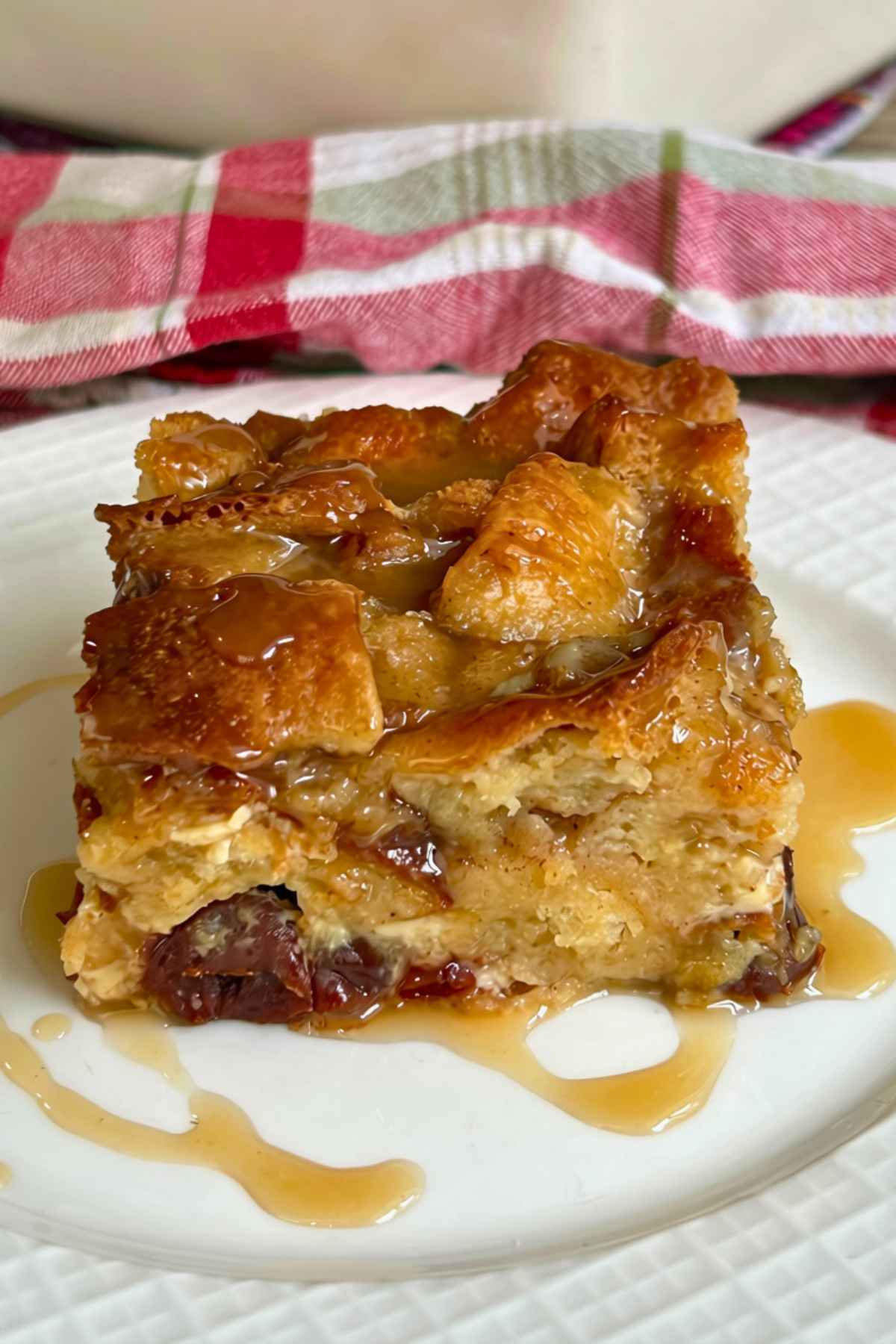 A square slice of warm old-fashioned bread pudding with raisins, drizzled with a butterscotch sauce, served on a white plate in front of a white baking dish on a red and white checkered cloth.