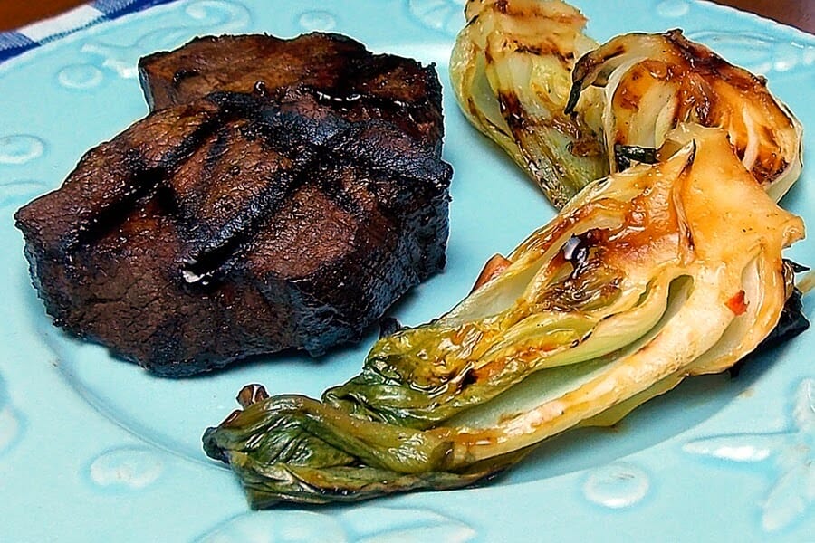 One marinated and grilled venison tenderloin steak with a side of grilled baby bok choy on a blue seashell plate.